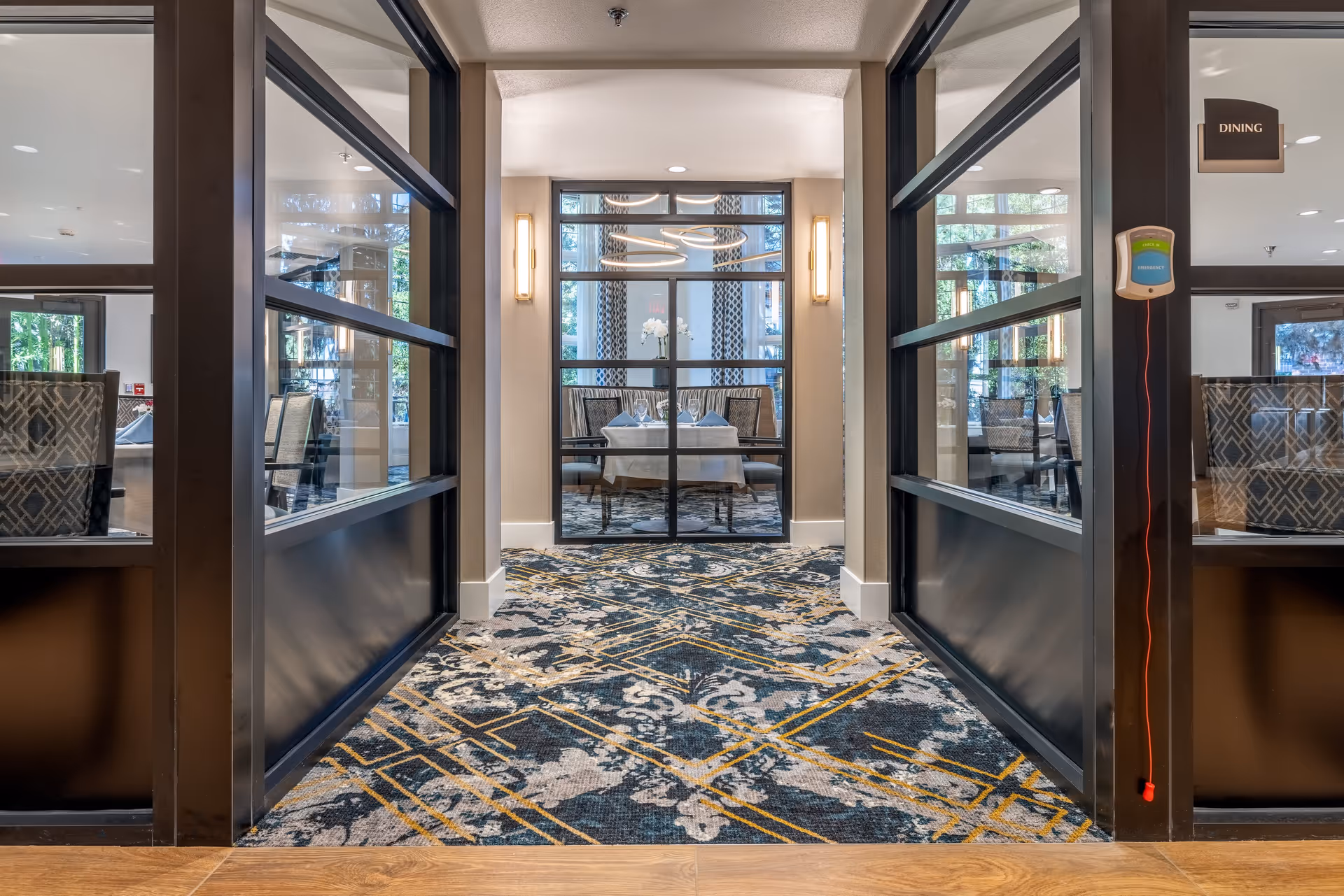 View down a hallway with patterned carpet leading to a dining area with a table set for four. The hallway is flanked by glass walls with black frames, and the dining area is illuminated by modern circular pendant lights. There are chairs with patterned upholstery visible through the glass walls.