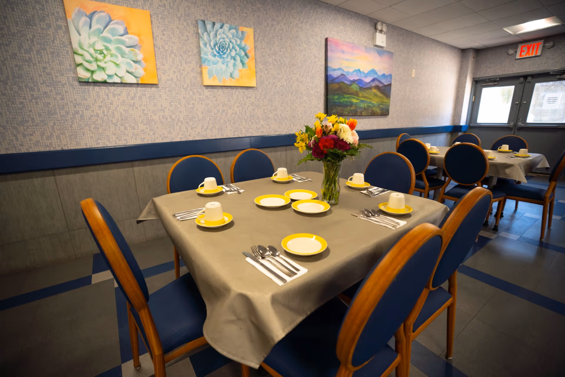 A dining room with tables covered in gray tablecloths, set with yellow-rimmed plates, cups, and silverware. Blue cushioned chairs with wooden frames surround the tables. A vase with colorful flowers is placed on the nearest table. The walls are decorated with paintings of flowers and a landscape, and there is an exit door in the background.