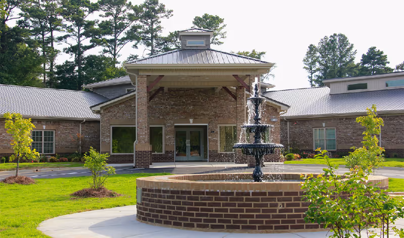 Front exterior view of a brick healthcare facility with a covered entrance and a multi-tiered water fountain in the foreground, surrounded by green grass, small trees, and shrubs.