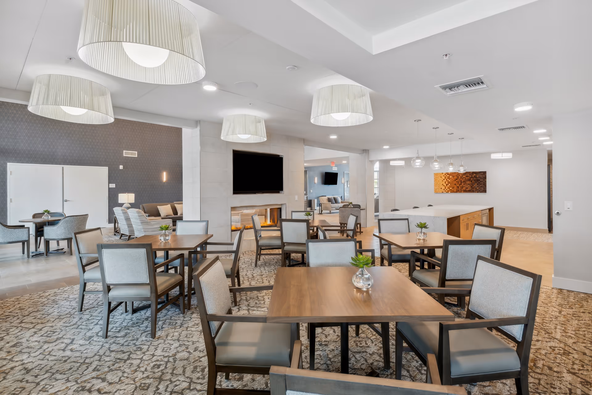 Bright communal dining and lounge area with multiple wooden tables and upholstered chairs, pendant lights, and a fireplace with a wall-mounted TV.