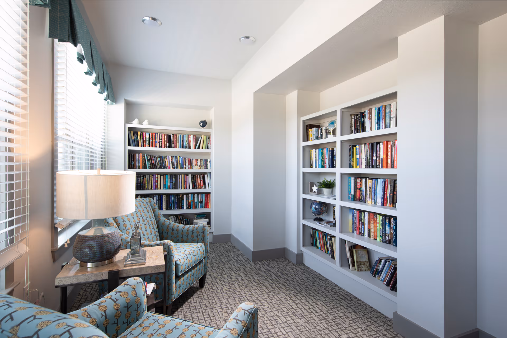 Bright reading nook with upholstered armchairs, a side table and lamp, and built-in bookshelves lining white walls.