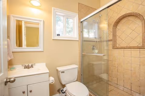 A small bathroom with beige walls featuring a white vanity with a sink and faucet, a mirror above the sink, a toilet next to the vanity, and a glass-enclosed shower with beige tiled walls and a built-in niche.