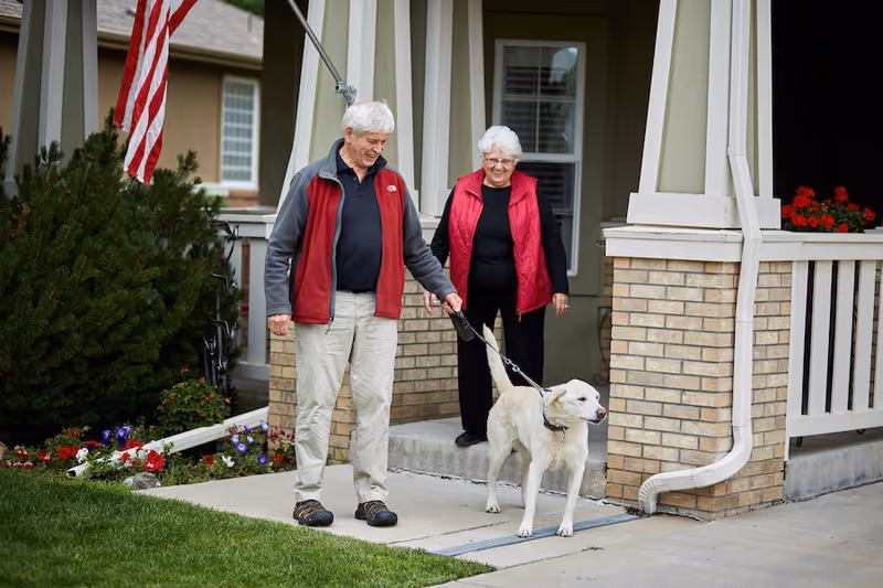 An elderly man and woman standing outside a house with brick pillars and a porch. The man is holding a leash and walking a white dog. There is an American flag hanging near the porch and flowers planted along the edge of the house.