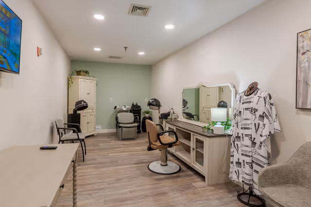 Interior view of a salon area in a senior living facility with salon chairs, hair drying stations, a large mirror, a mannequin wearing a barber cape, and a small table with a lamp and plant. The room has light-colored walls and wood flooring.