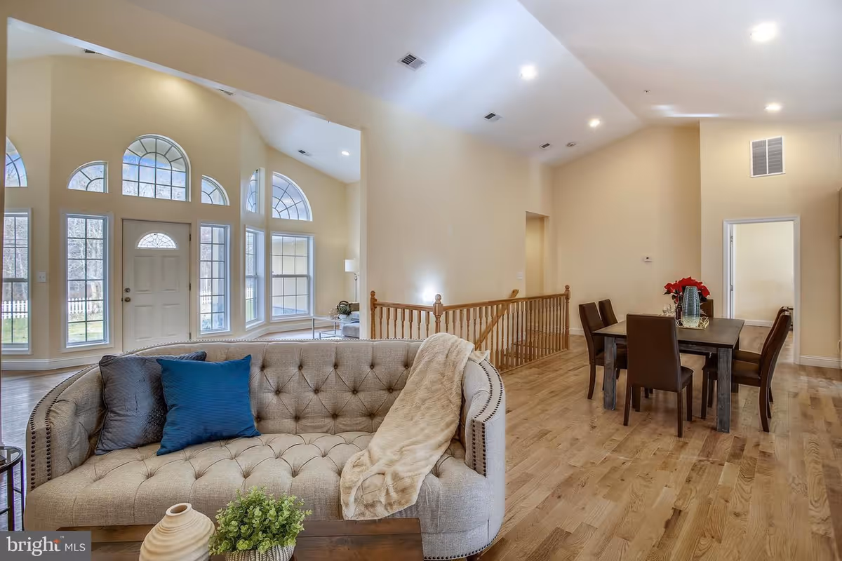 Interior view of a senior living facility showing a spacious living room and dining area. The living room features a tufted beige sofa with blue and gray pillows and a beige throw blanket. There is a wooden coffee table with a small plant and decorative items. Large arched windows and a door let in natural light. The dining area has a dark wooden table with six chairs and a vase with red flowers. The floor is light hardwood, and the walls are painted a soft beige color.
