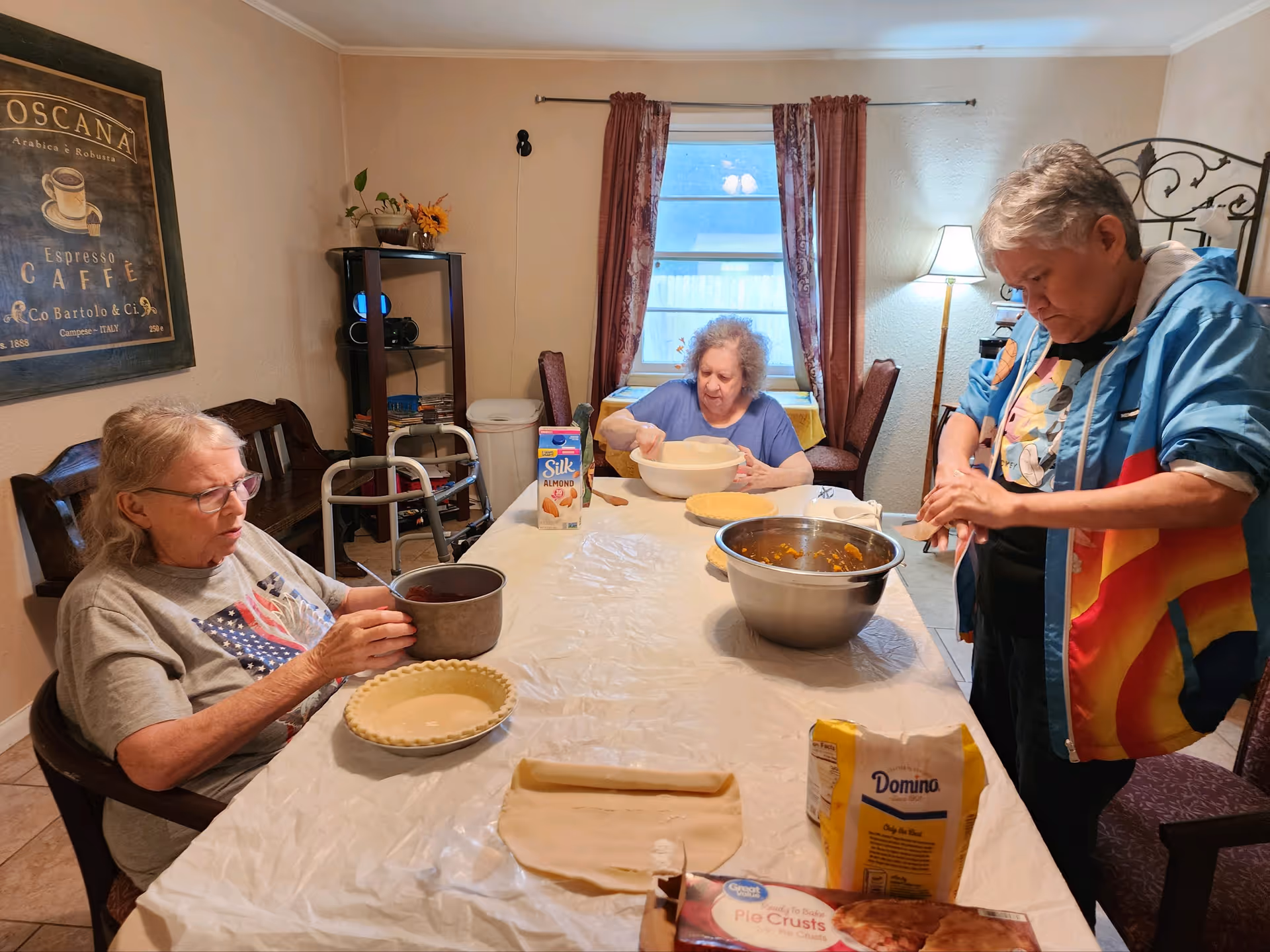 Three elderly women in a dining room preparing pie crusts and fillings on a long table covered with a white tablecloth. Various baking ingredients and utensils are on the table, including pie crusts, a carton of almond milk, a bag of sugar, and mixing bowls. The room has a window with curtains, a floor lamp, and a framed coffee-themed picture on the wall.