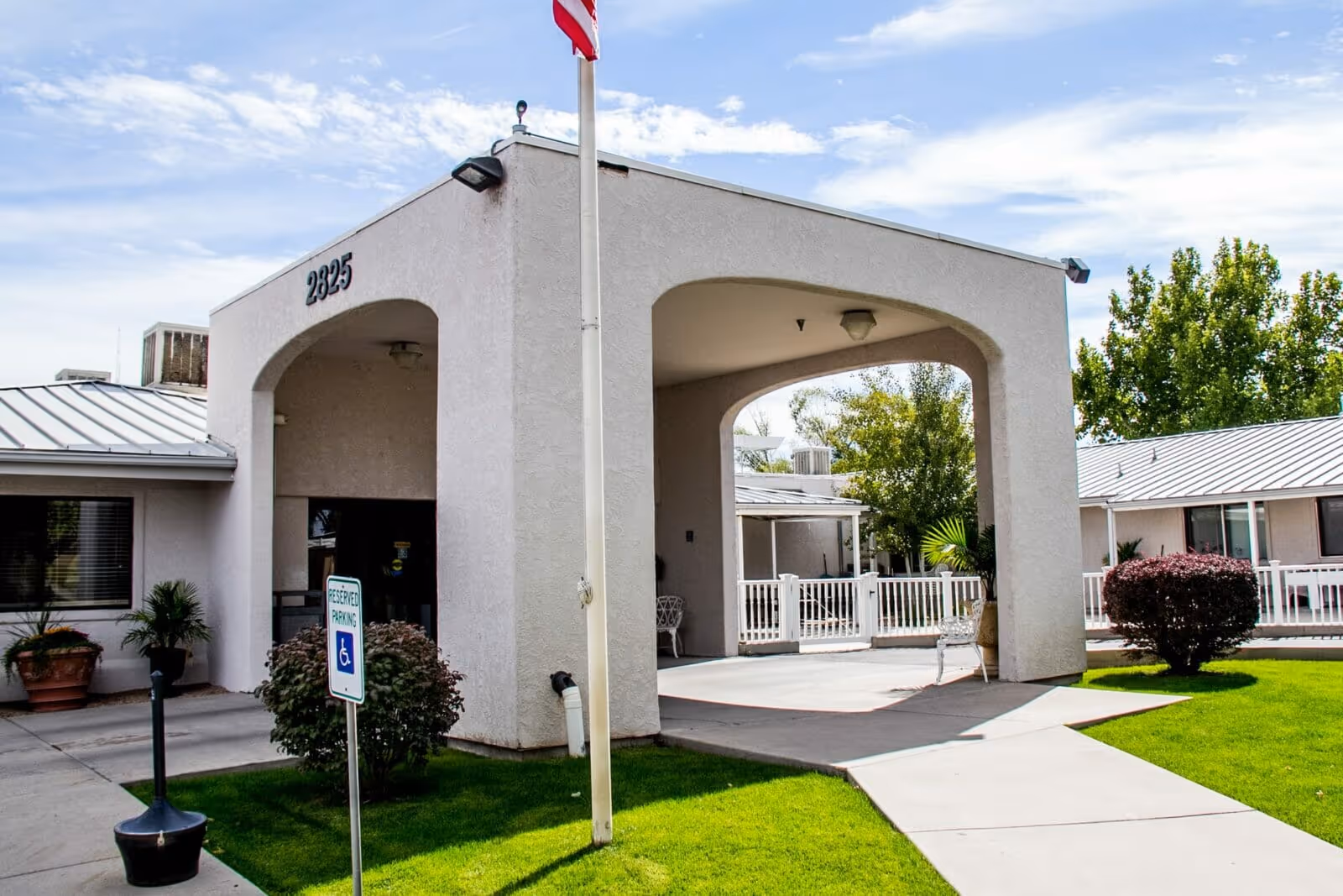 Front entrance canopy of a single-story care facility showing the address number '2825', a flagpole, accessible parking sign, and landscaped lawn.