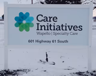 Outdoor sign reading "Care Initiatives Wapello | Specialty Care, 601 Highway 61 South" standing on a snowy lawn.