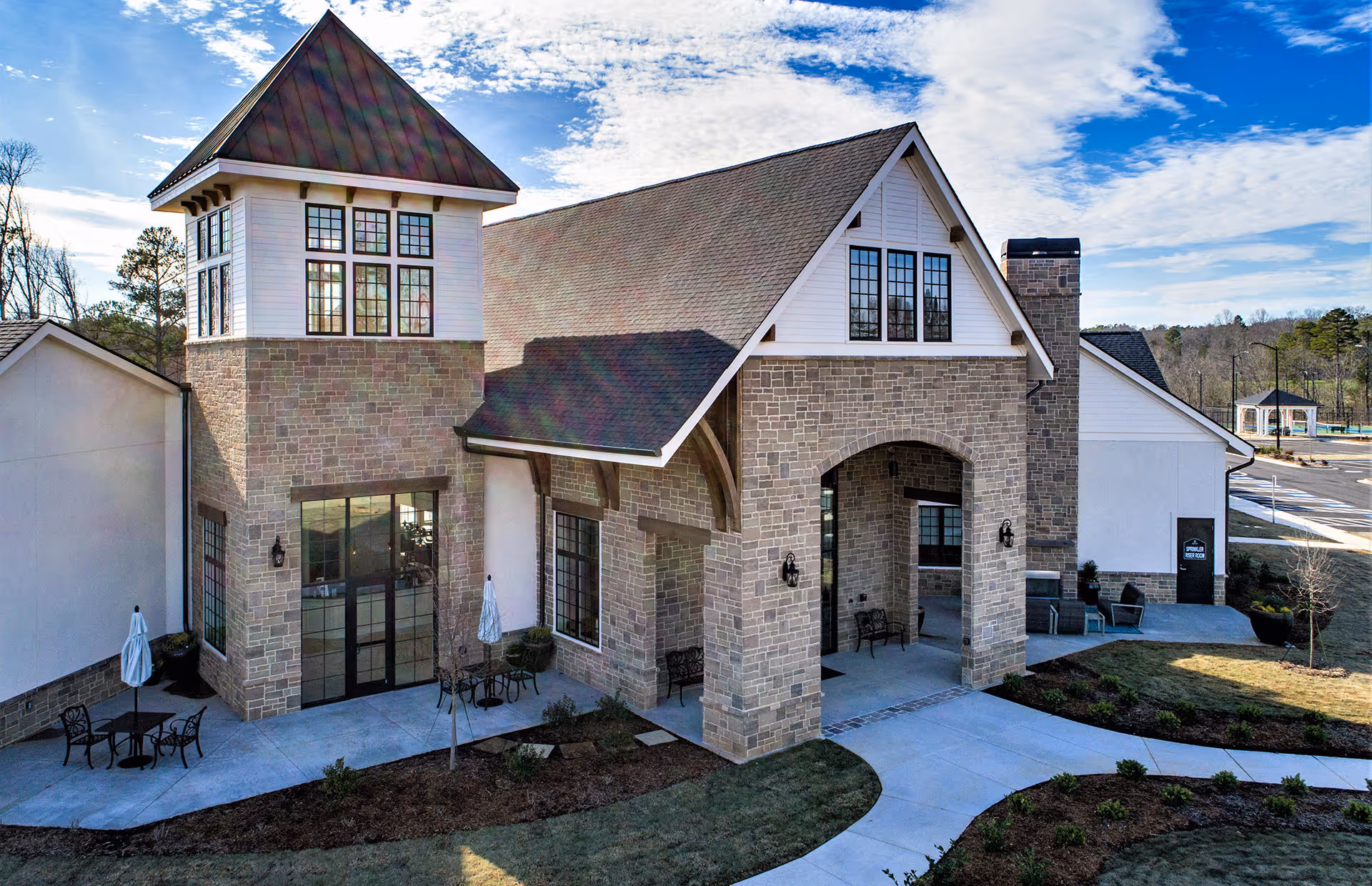 Stone-and-white siding clubhouse with an arched covered entrance, patio seating, and landscaped walkways under a blue sky.