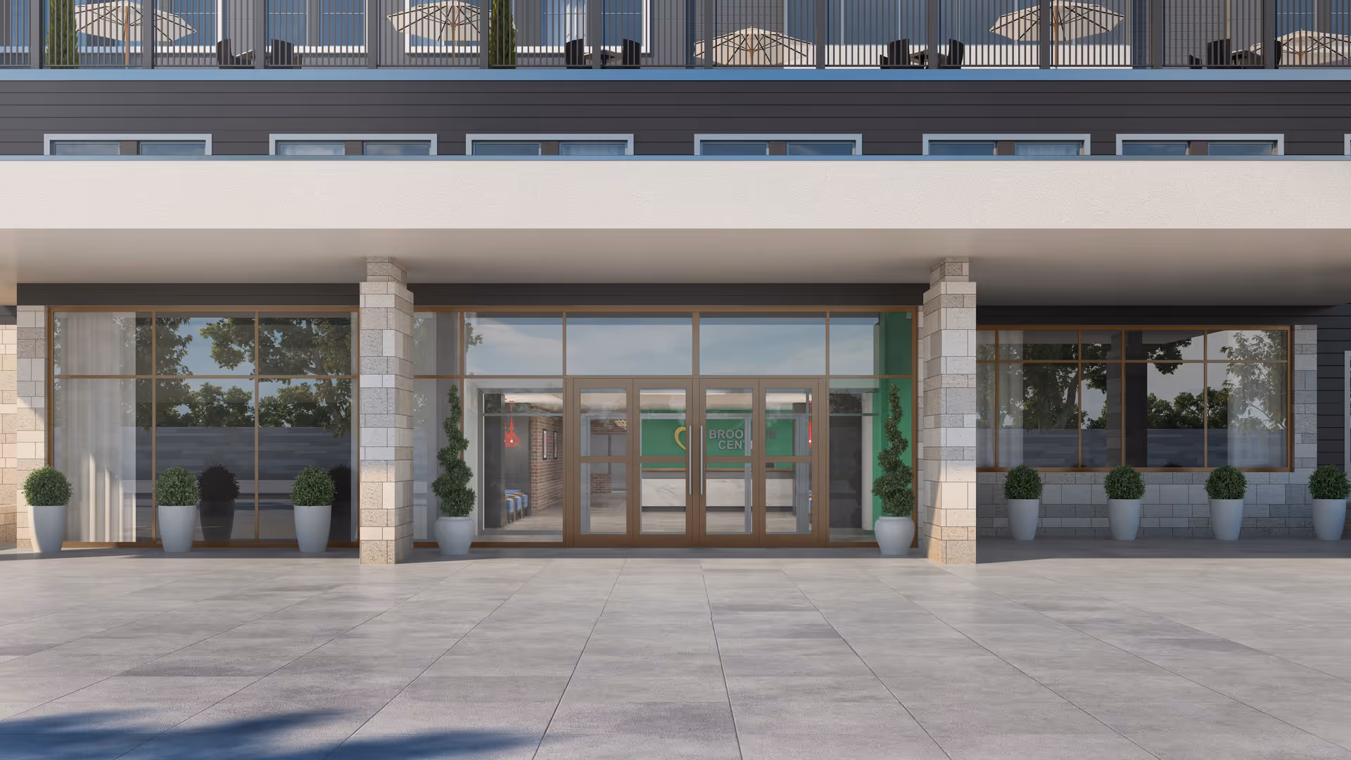 Front exterior view of a modern building entrance with large glass doors and windows, flanked by potted plants. The building has a covered entryway supported by stone pillars and a balcony with railings above.