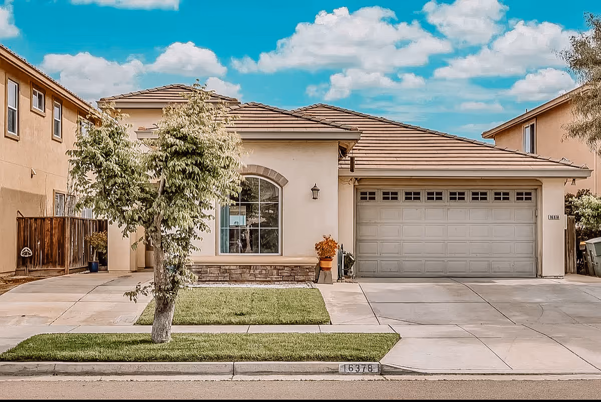 Front exterior view of a single-story house with a tiled roof, a large garage door, a window with an arched top, a small tree in the front yard, and a clear blue sky with some clouds.