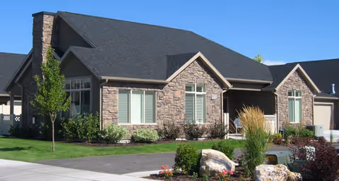 Single-story residential building with stone facade, black roof, and multiple windows with white shutters. The house is surrounded by a well-maintained lawn, small trees, shrubs, and decorative rocks under a clear blue sky.