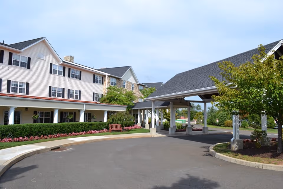 Front exterior of a multi-story senior living building with a covered porte-cochere, driveway, benches, and landscaped flowerbeds.