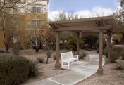 Outdoor courtyard with a wooden pergola and white bench along a paved path surrounded by desert landscaping and a beige building.