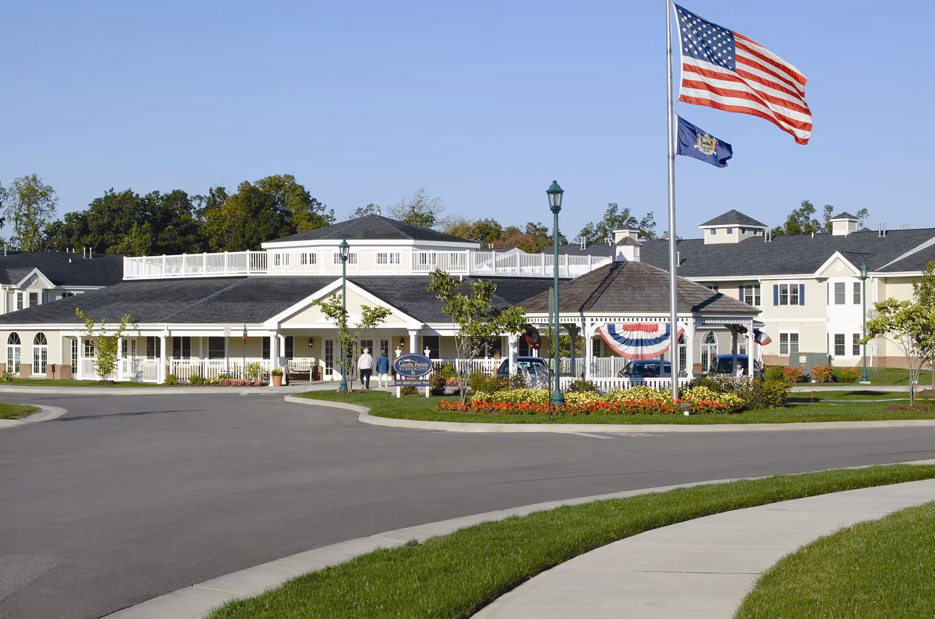 Exterior view of a senior living facility with a circular driveway, landscaped flower beds, and two flagpoles displaying the American flag and another flag. The building is light-colored with a dark roof and features a covered entrance and a gazebo decorated with patriotic bunting.