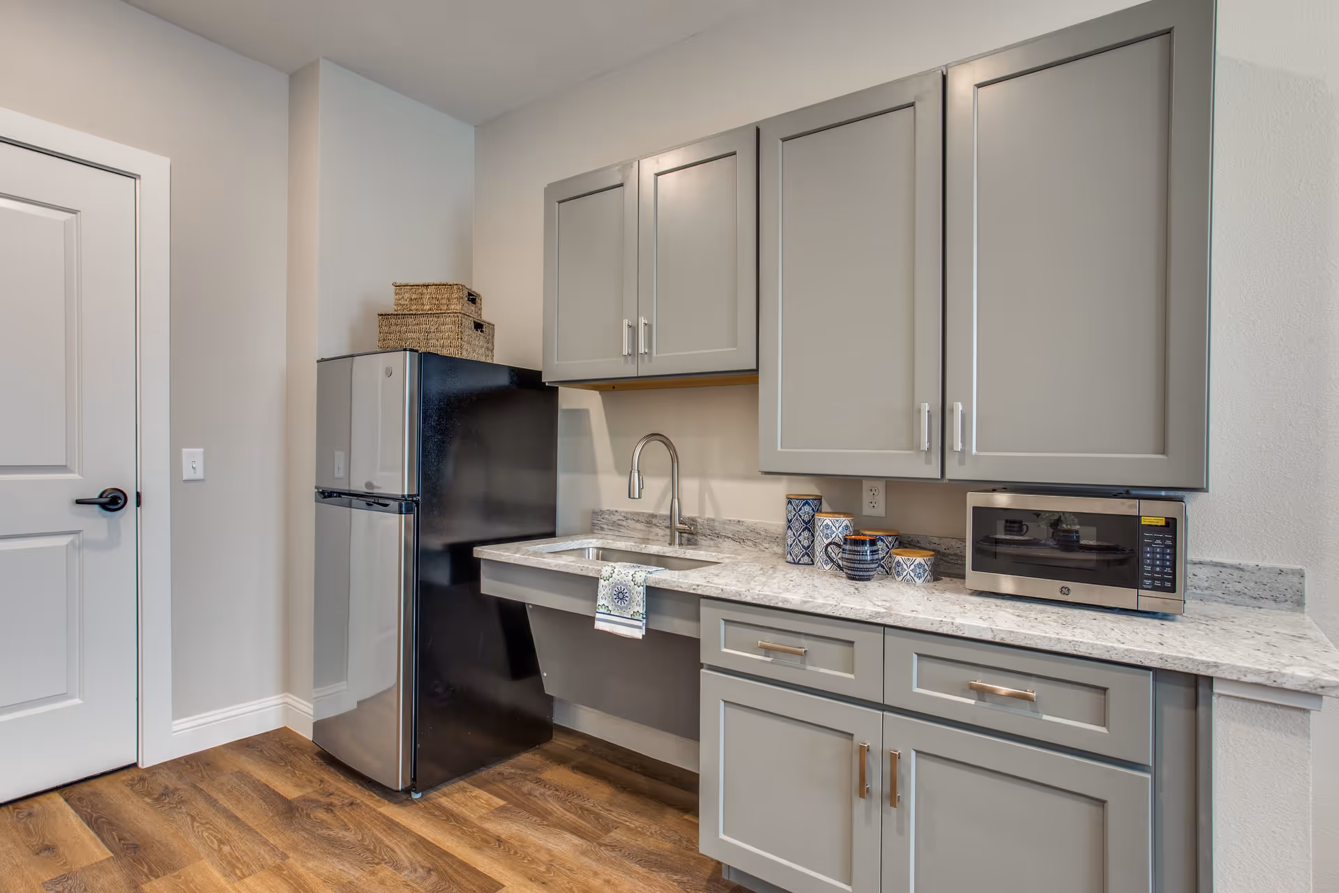 Small kitchenette with gray cabinets, marble countertop, sink, microwave, and a black refrigerator.