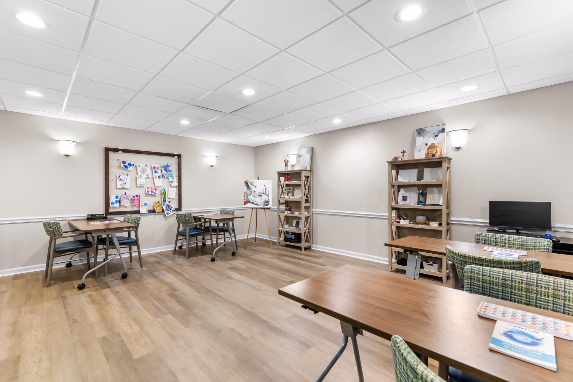 Bright interior activity room with tables and chairs, shelving, a bulletin board of artwork, and a small TV.