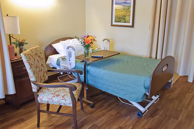 A hospital-style bed with a green bedspread and white pillows in a room with wooden flooring. Next to the bed is a wooden nightstand with a lamp and a vase of flowers. A floral upholstered armchair is positioned near the bed, and a small overbed table with a vase of flowers and a remote control is placed over the bed. A framed landscape picture hangs on the wall above the bed, and beige curtains cover the window.