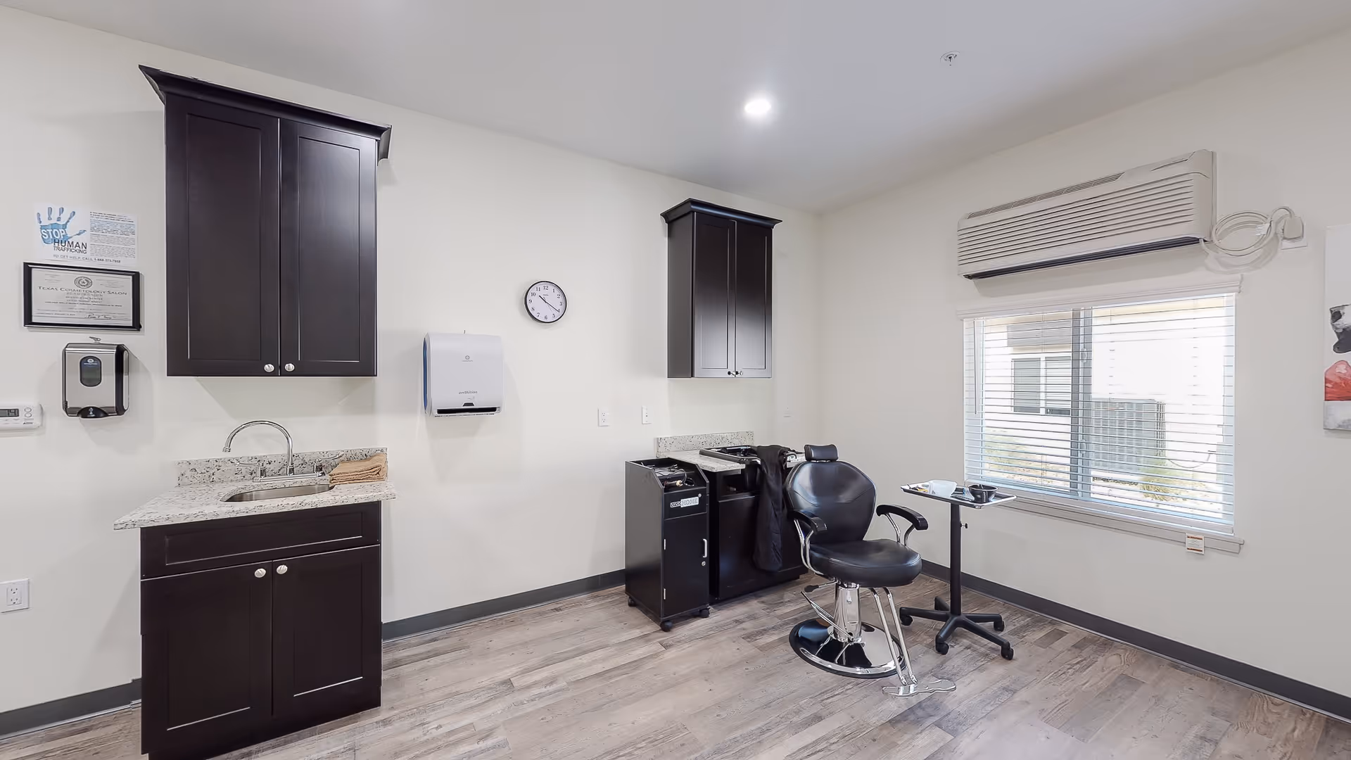 A clean and modern salon room with dark wood cabinets, a small sink with countertop, a black salon chair, a rolling cart, a small table with hairdressing tools, a wall clock, and a window with blinds. The walls are light-colored and the floor has a wood-like finish.