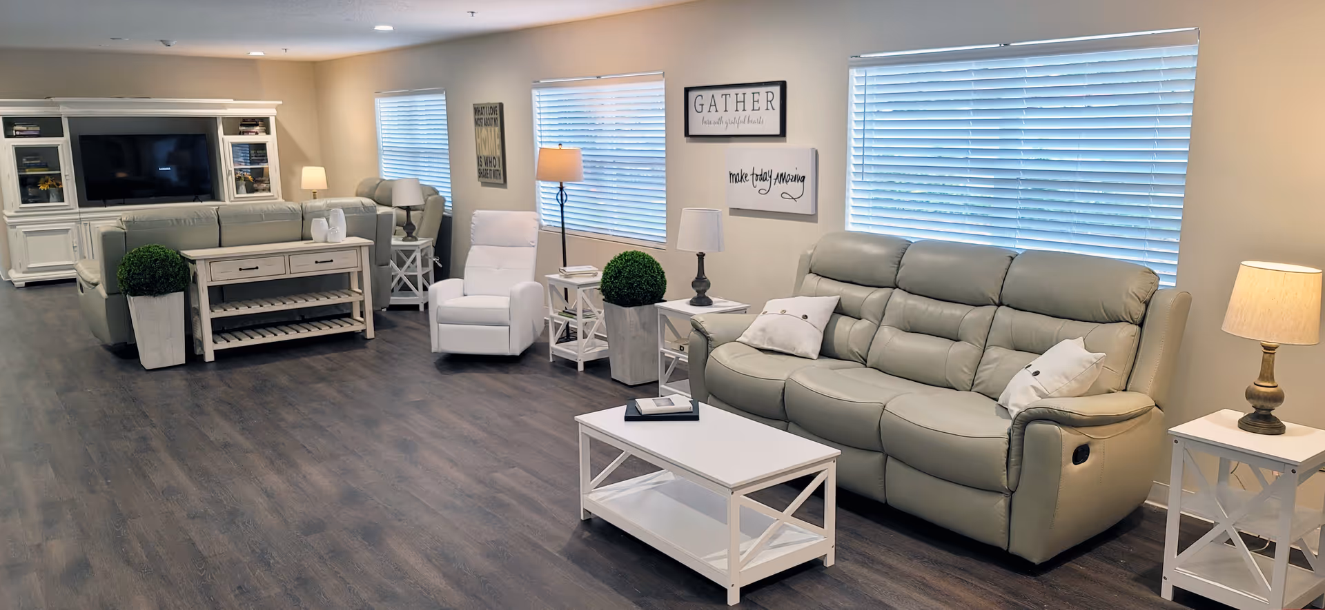 Bright common living area with beige recliner sofas, white accent tables, lamps, and a large TV cabinet.