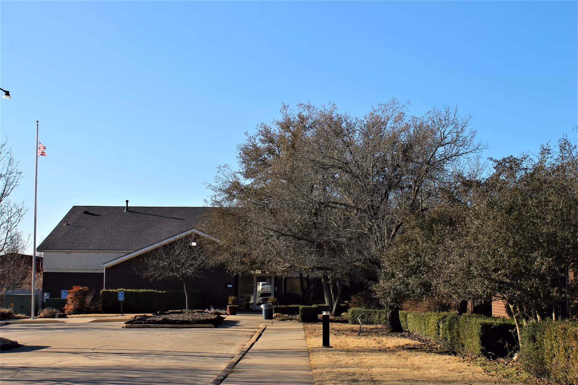 Exterior view of Brentwood Nursing Home, Extended Care and Rehabilitation building with a clear blue sky, leafless trees, a flagpole with an American flag, and a paved driveway leading to the entrance.