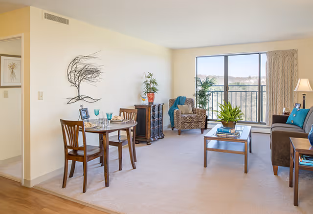 A bright living room and dining area in an apartment with a small round dining table set for two, a patterned armchair, a brown sofa with blue cushions, a wooden coffee table with a plant, and a large window with a view outside.