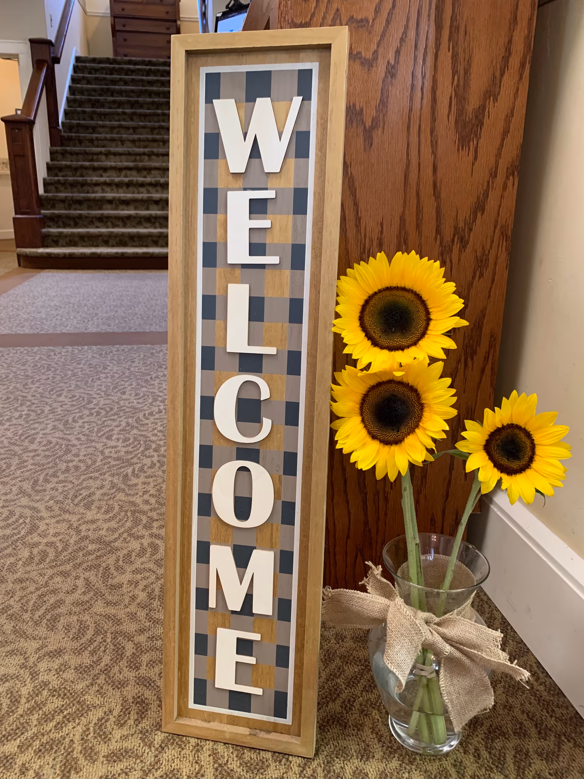 A vertical wooden sign with the word 'WELCOME' in large white letters on a checkered background, placed on a carpeted floor next to a wooden wall panel. Beside the sign is a clear glass vase with three bright yellow sunflowers tied with a burlap ribbon. In the background, there is a carpeted staircase with wooden handrails.