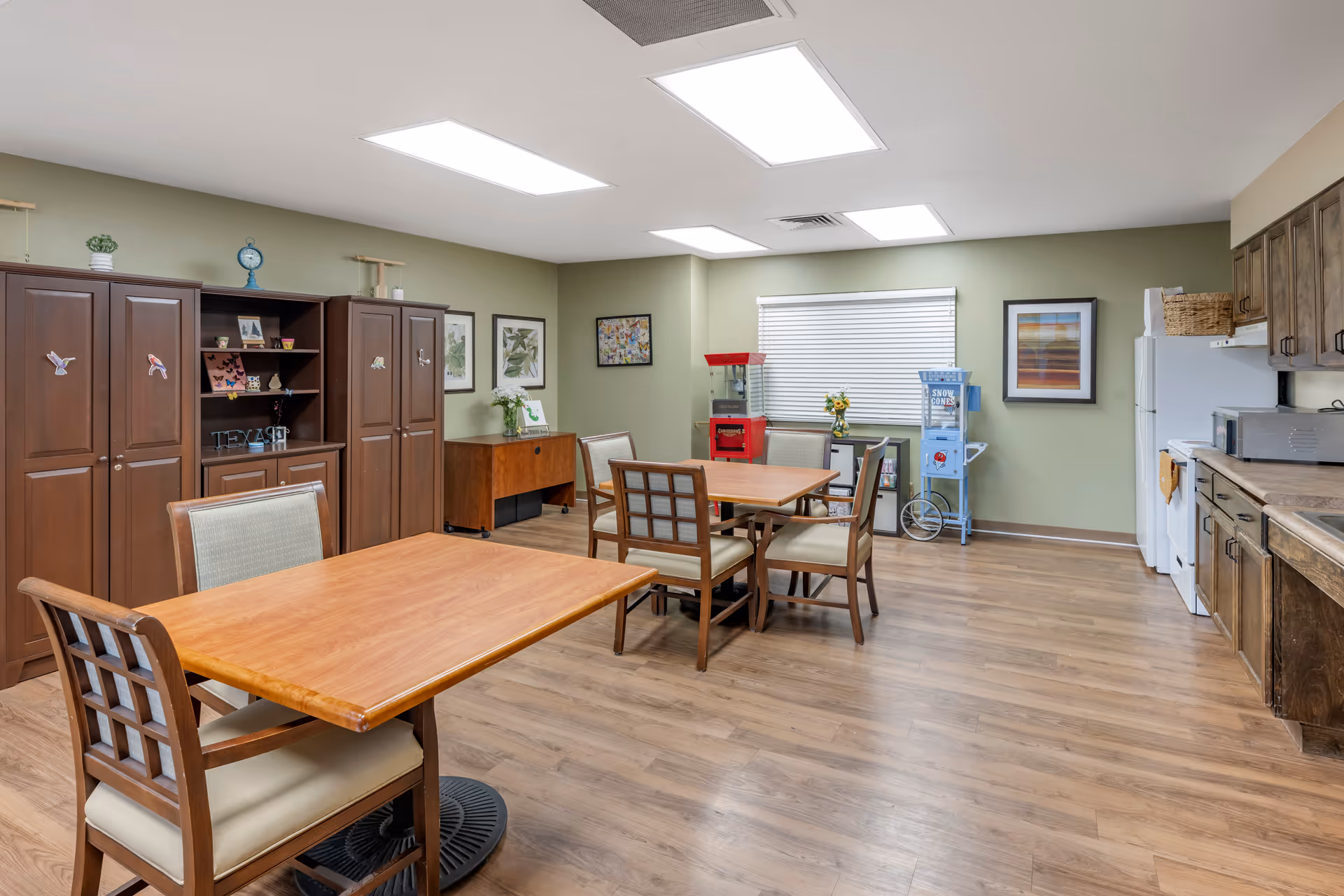 A spacious room with wooden flooring and light green walls featuring two wooden tables with chairs around them. On the right side, there is a kitchenette with wooden cabinets, a white refrigerator, microwave, and stove. The back wall has a window with blinds, a red popcorn machine, a blue snow cone machine, and framed artwork. On the left side, there are tall wooden cabinets and shelves decorated with small plants, pictures, and ornaments.