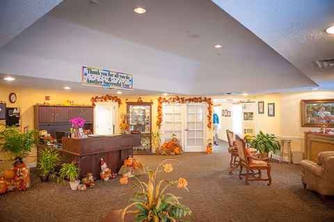 Interior view of a senior living facility lobby or common area decorated with autumn-themed decorations including pumpkins and garlands. There is a wooden reception desk with plants and flowers on it, comfortable seating with chairs and a sofa, and a glass display cabinet. A banner hanging from the ceiling reads 'Thank you for all you do & Another Successful Year!'. The space is warmly lit and welcoming.