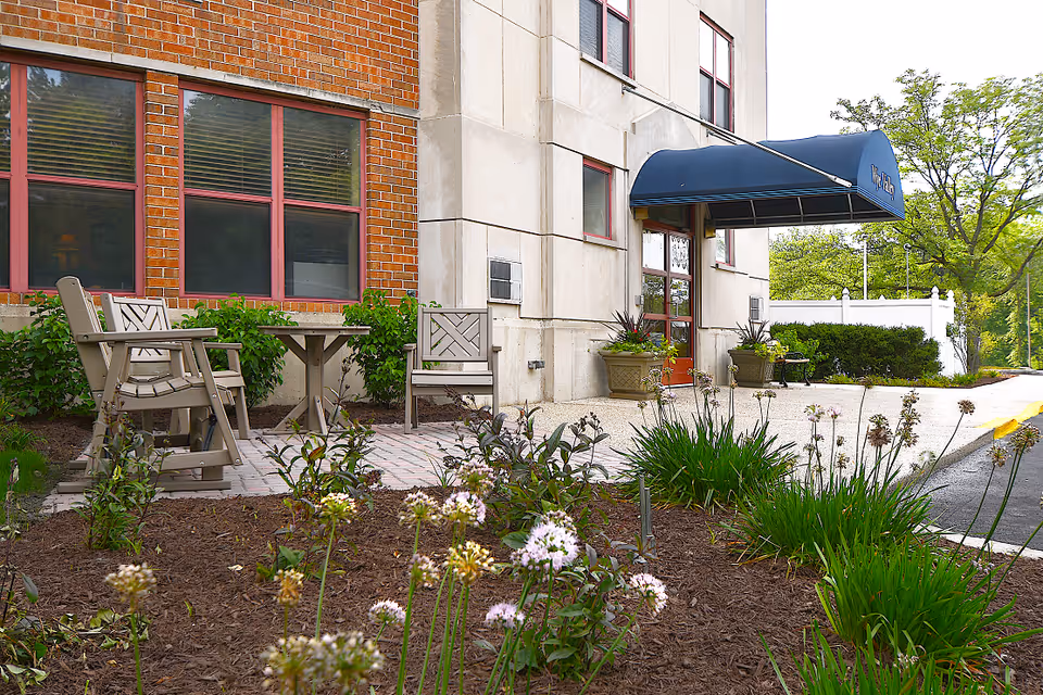 Outdoor seating area with two chairs and a table on a paved patio next to a building entrance with a blue awning. There are plants and flowers in the foreground and bushes along the side of the building.