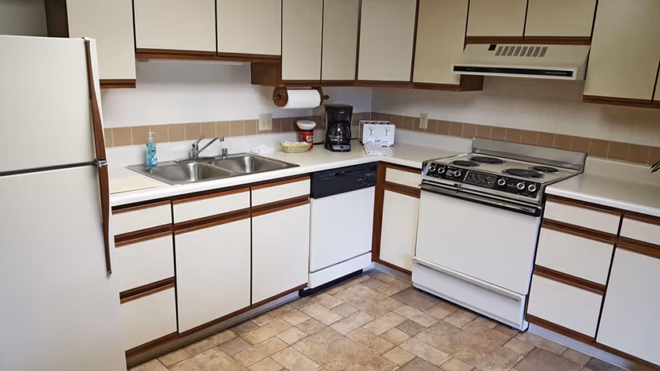 A kitchen area with white cabinets featuring wooden handles, a white refrigerator, a double sink with a soap dispenser, a dishwasher, a white electric stove with an oven, a coffee maker, and a toaster on the countertop. The floor is tiled with a beige and brown pattern.