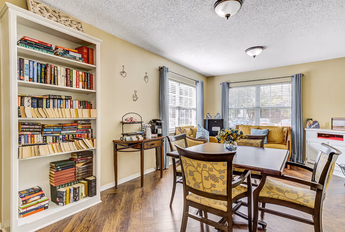 A cozy common area with a wooden table and four upholstered chairs featuring a floral pattern. Behind the table is a yellow cushioned bench with blue pillows, positioned in front of two large windows with gray curtains. To the left, there is a tall white bookshelf filled with books and a small wooden table with a coffee maker and cups. The room has wooden flooring and beige walls with simple wall decorations.