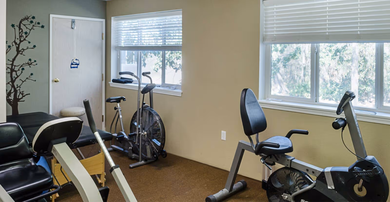 Small interior exercise room with stationary and recumbent bikes, a window, and beige walls.