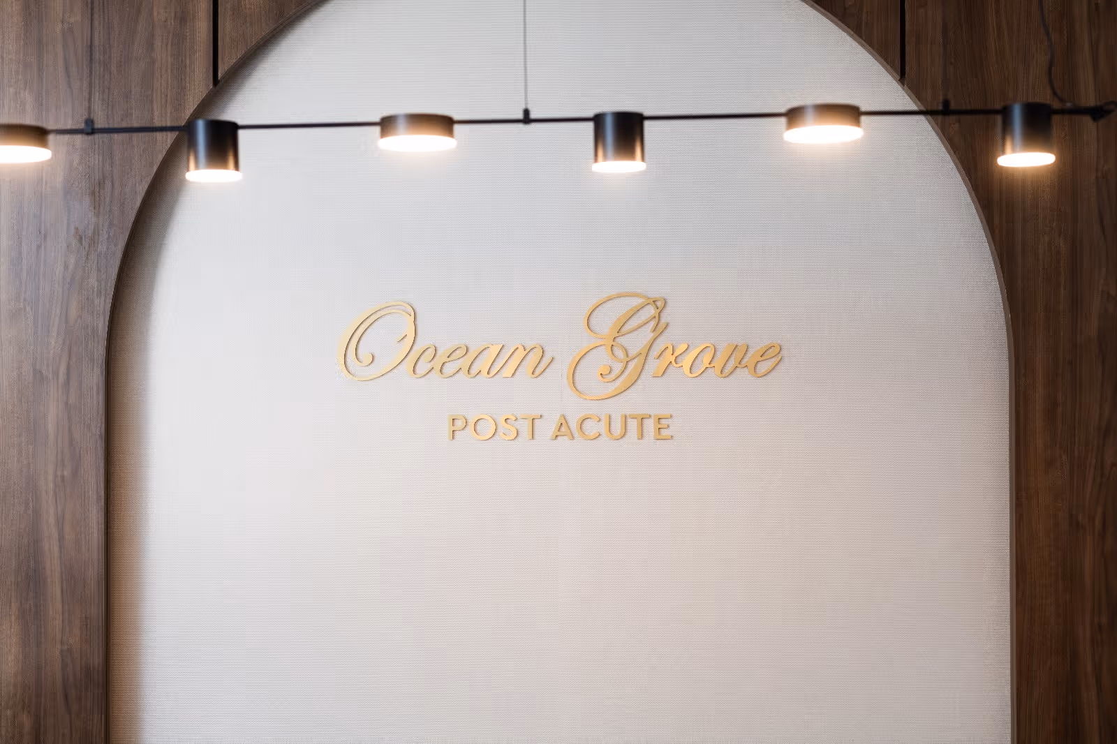 A wall with a large arch design featuring the text 'Ocean Grove POST ACUTE' in gold lettering, illuminated by a row of modern black ceiling lights.