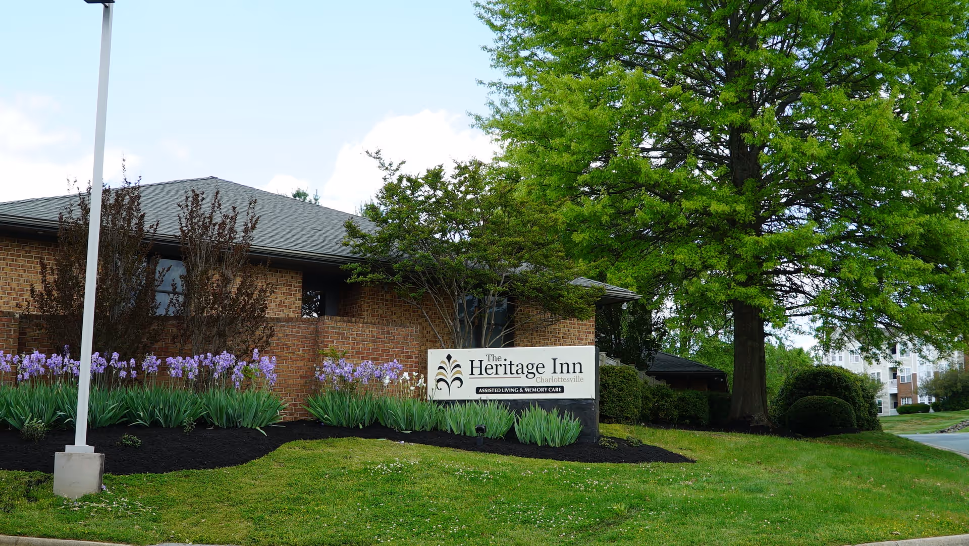 Exterior view of The Heritage Inn Assisted Living and Memory Care building with a brick facade, surrounded by green grass, purple flowers, bushes, and a large tree. A sign in front displays the facility's name.