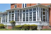 A white enclosed sunroom with tall multi-pane windows along the ground floor of a red brick building with shrubs in front.