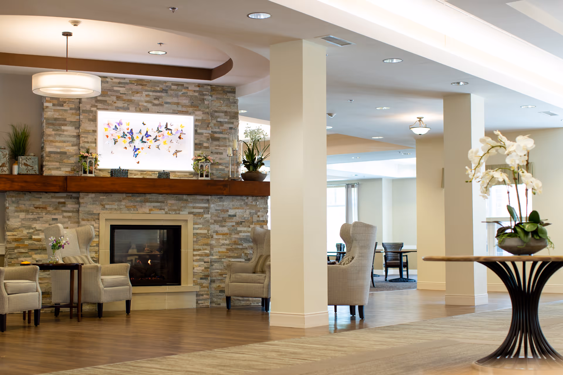 A spacious and well-lit common area with a stone fireplace, beige armchairs, a wooden mantel decorated with plants and small items, and a colorful butterfly artwork above the fireplace. The room has wooden flooring, light-colored walls, and a round table with a white orchid plant in the foreground.
