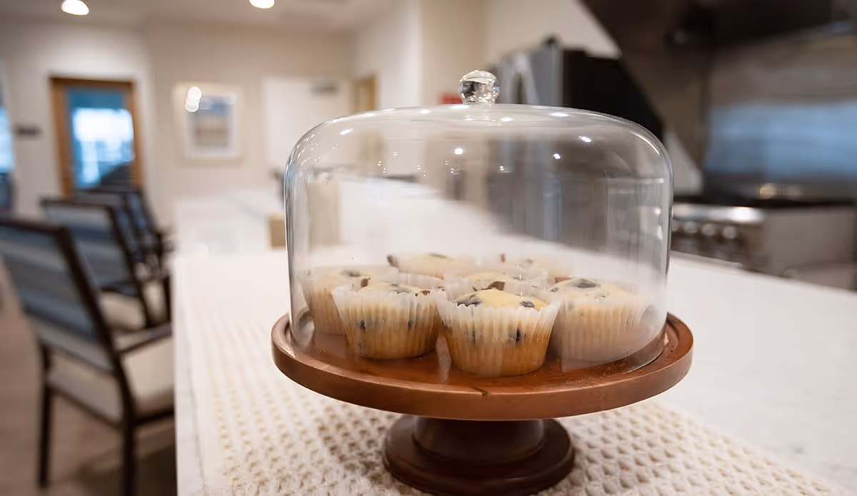 A close-up view of a wooden cake stand with a glass dome cover containing several blueberry muffins, placed on a white textured table runner in a kitchen setting with chairs and kitchen appliances in the background.