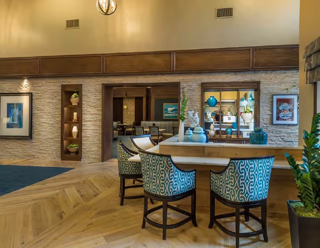 Interior view of a senior living facility lounge area with a wooden counter and three patterned upholstered chairs. The background features a stone accent wall with built-in shelves holding decorative items, framed artwork, and a window opening into another room with more seating and decor.
