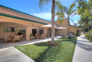Outdoor patio area at Summerfield of Encinitas Memory Care with wooden tables and chairs under a covered walkway, palm trees, umbrellas, lounge chairs, and a well-maintained lawn and sidewalk under a clear blue sky.