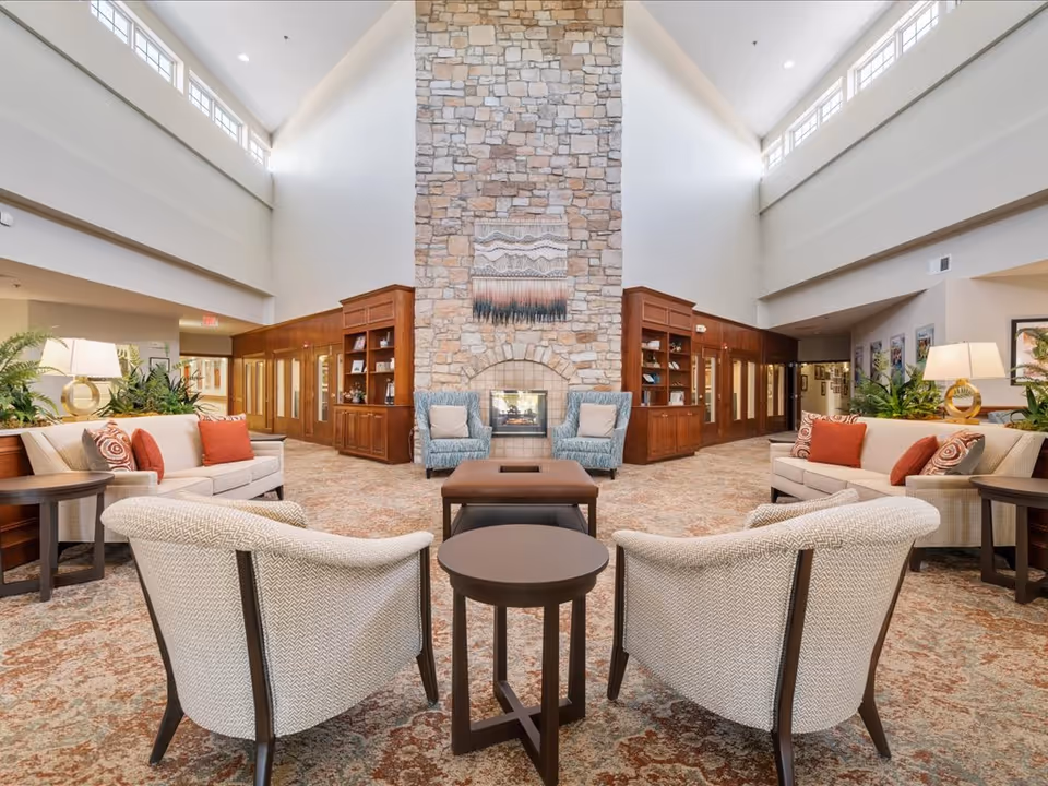 A spacious, well-lit lounge with high ceilings, a tall stone fireplace, sofas and armchairs arranged around a central coffee table and built-in wooden shelving.