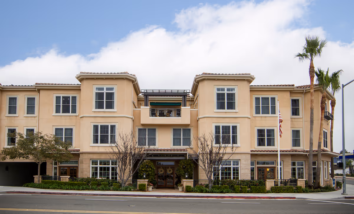 Front exterior view of a three-story beige building with multiple windows, a central entrance with greenery on either side, and palm trees on the right side. The building has a tiled roof and a balcony above the entrance. The sky is partly cloudy.