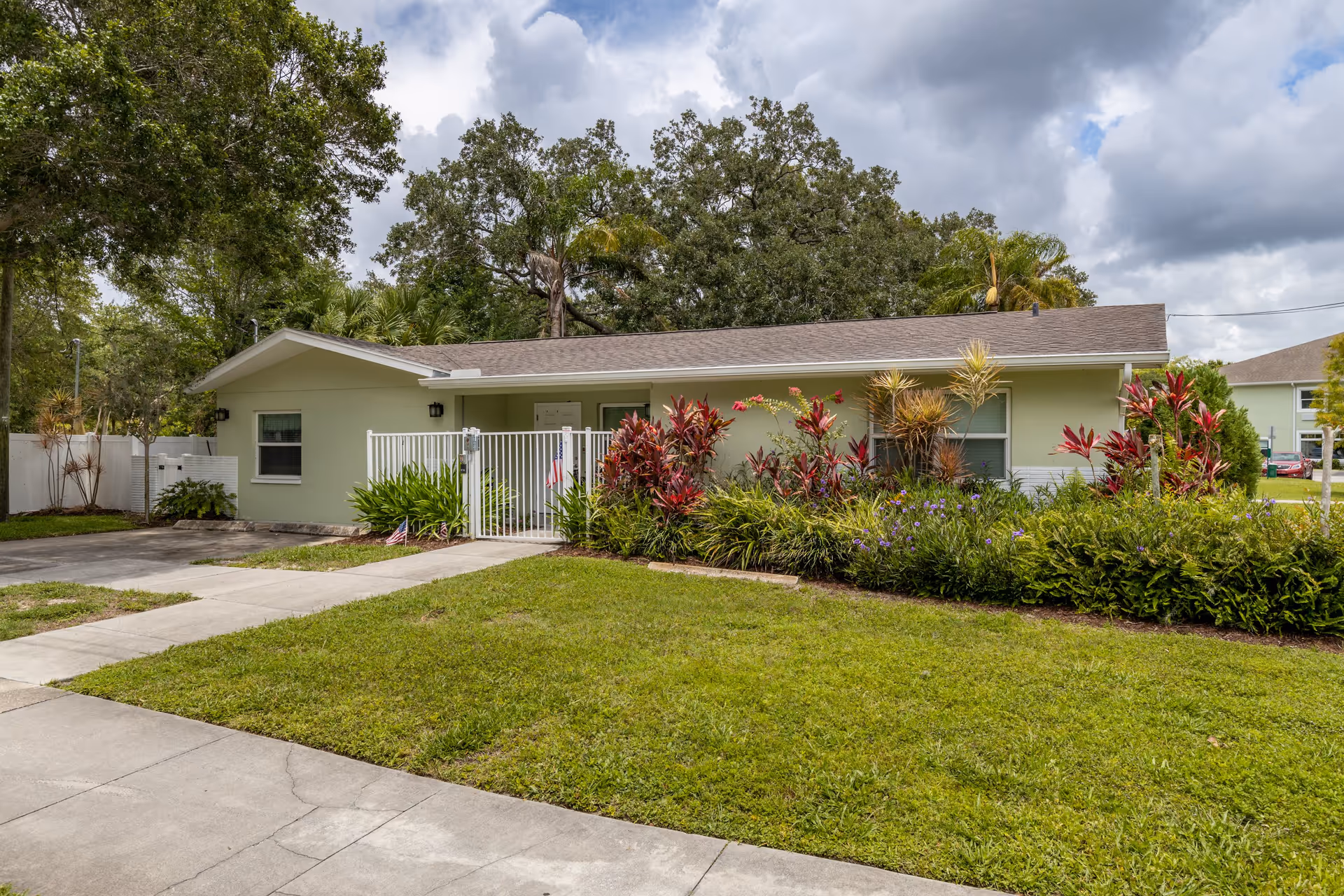 Single-story light green house with a brown shingled roof, surrounded by lush greenery and colorful plants. A white metal gate is in front of the entrance, and there is a concrete driveway and sidewalk leading up to the house. The sky is partly cloudy.
