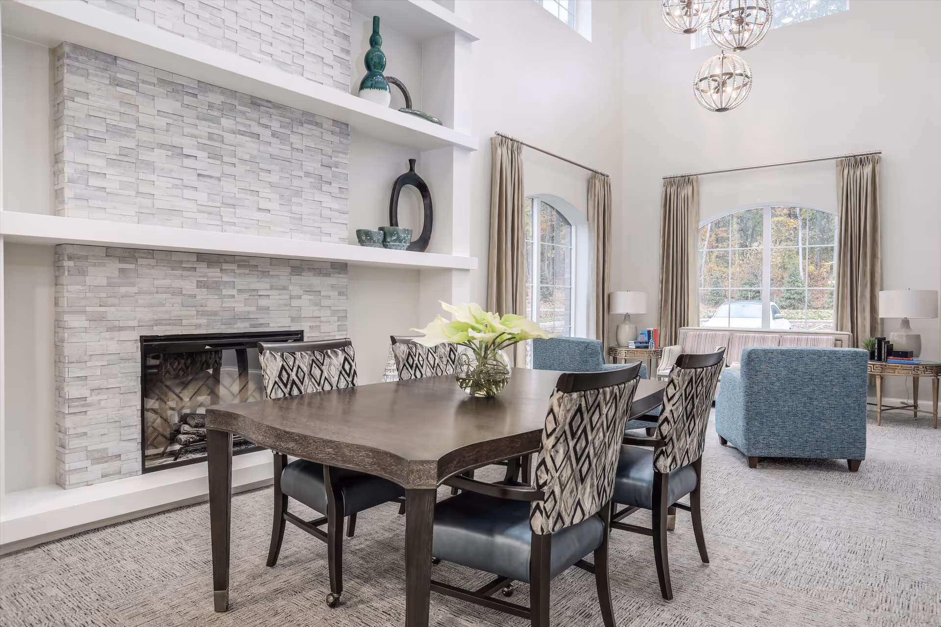 A bright and modern living room and dining area with a dark wooden dining table surrounded by six chairs, a stone fireplace with decorative shelves above it, large arched windows with beige curtains, two blue armchairs, a beige sofa, side tables with lamps, and three spherical pendant lights hanging from the ceiling.