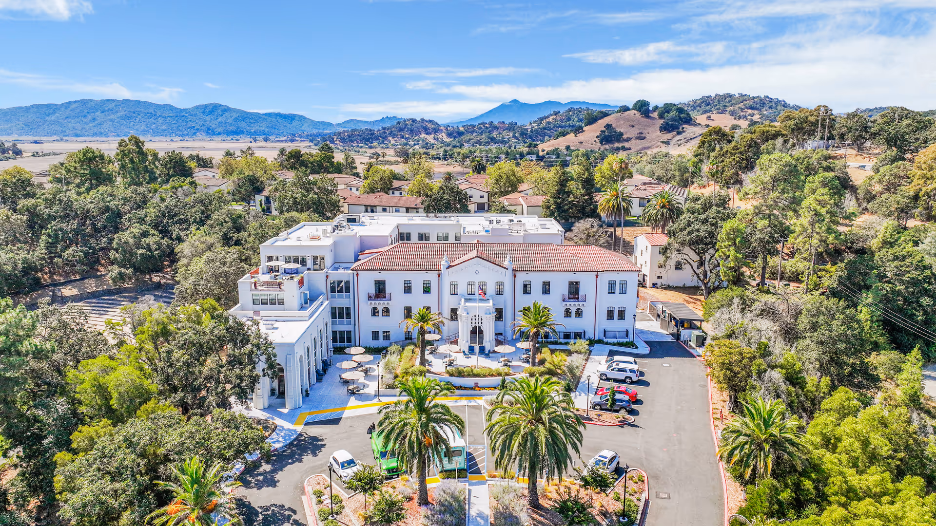 Aerial view of The Bluffs at Hamilton Hill facility surrounded by trees and hills. The building has a white exterior with a red-tiled roof and an outdoor seating area with tables and umbrellas in front. Several cars are parked in the parking lot, and the landscape includes palm trees and greenery.
