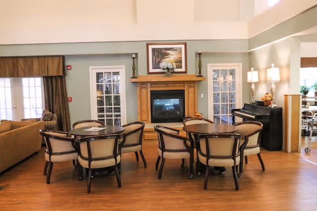 Communal lounge with round tables and chairs arranged before a fireplace, piano, and seating area.
