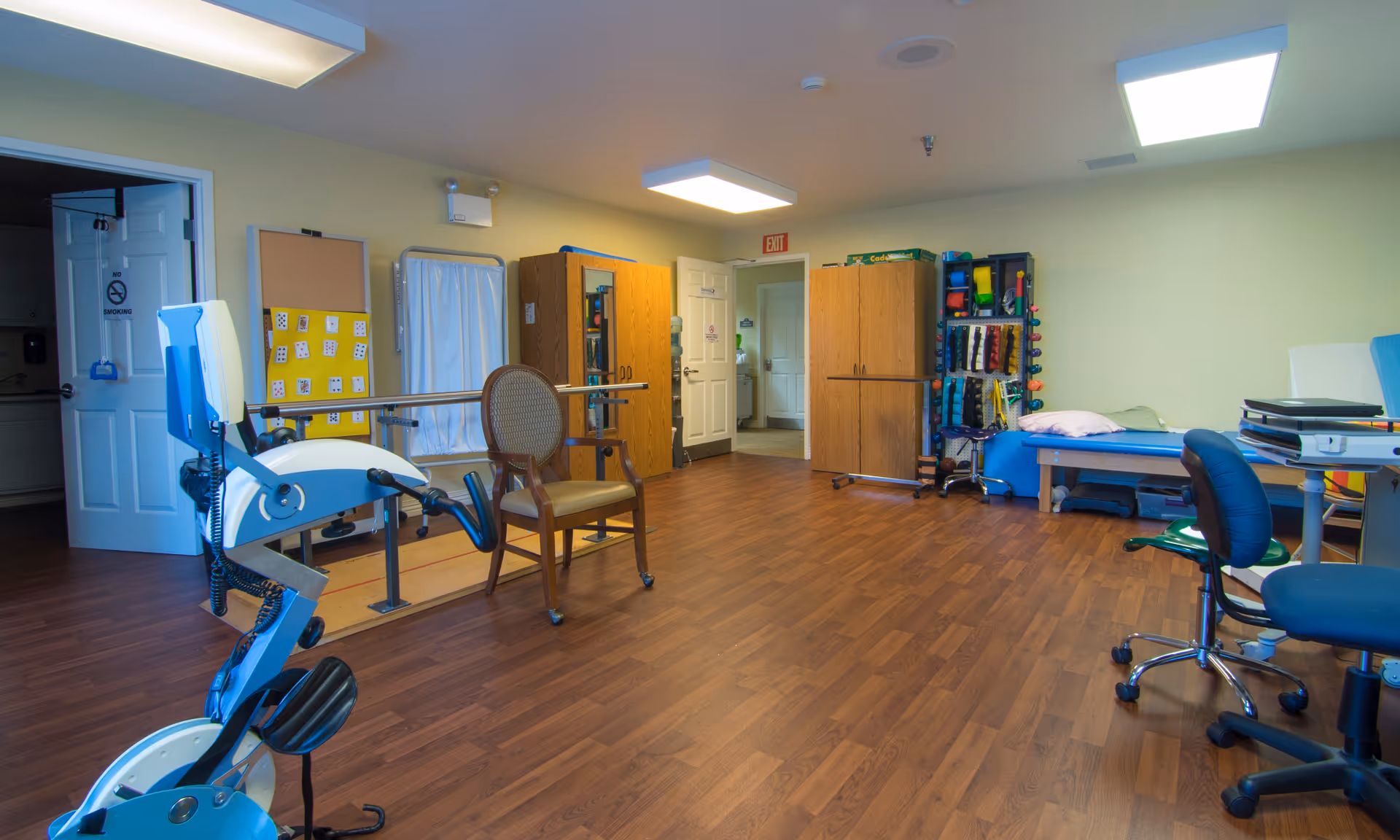 A therapy or rehabilitation room with wooden flooring, exercise equipment including a stationary bike, parallel bars with a chair between them, storage cabinets, and a therapy table with pillows. The room has fluorescent ceiling lights and an open door leading to another area.