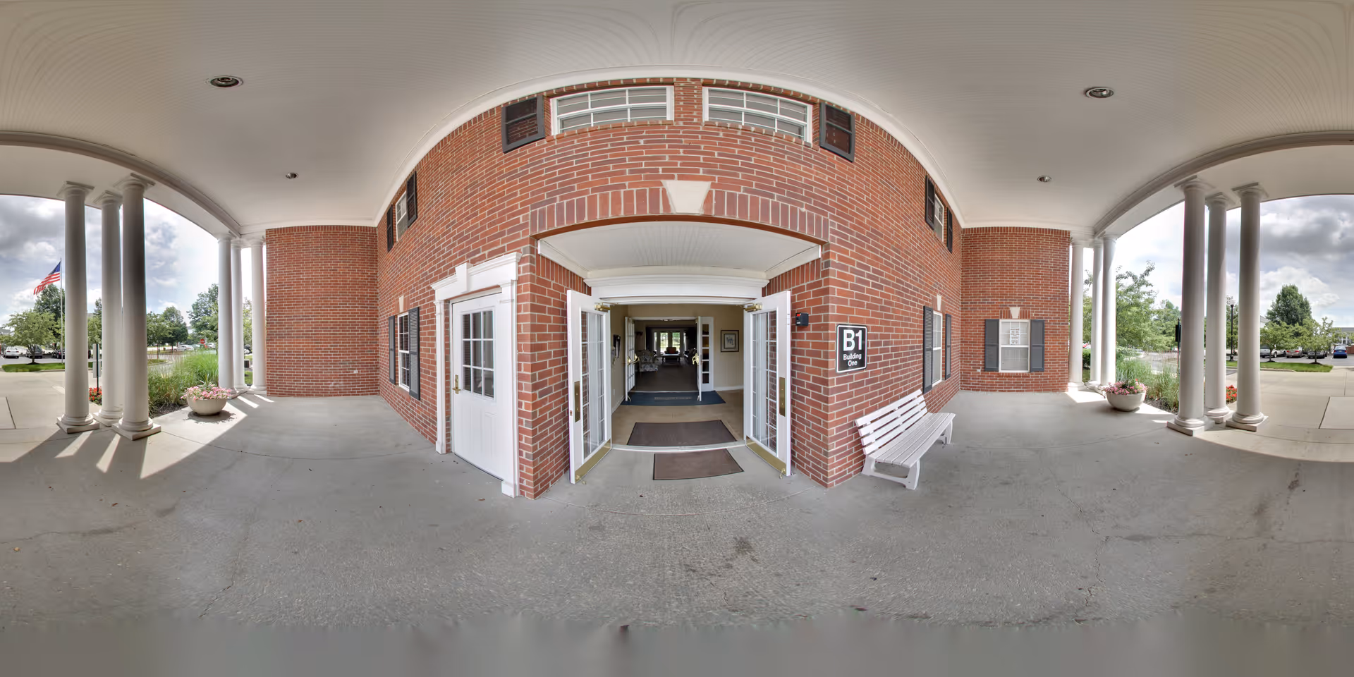 Covered entrance area of a brick building with white columns and double glass doors leading inside. There is a white bench on the right side and potted plants on both sides near the columns. A sign next to the door reads 'B1 Building One'. Trees and a parking lot are visible in the background under a partly cloudy sky.