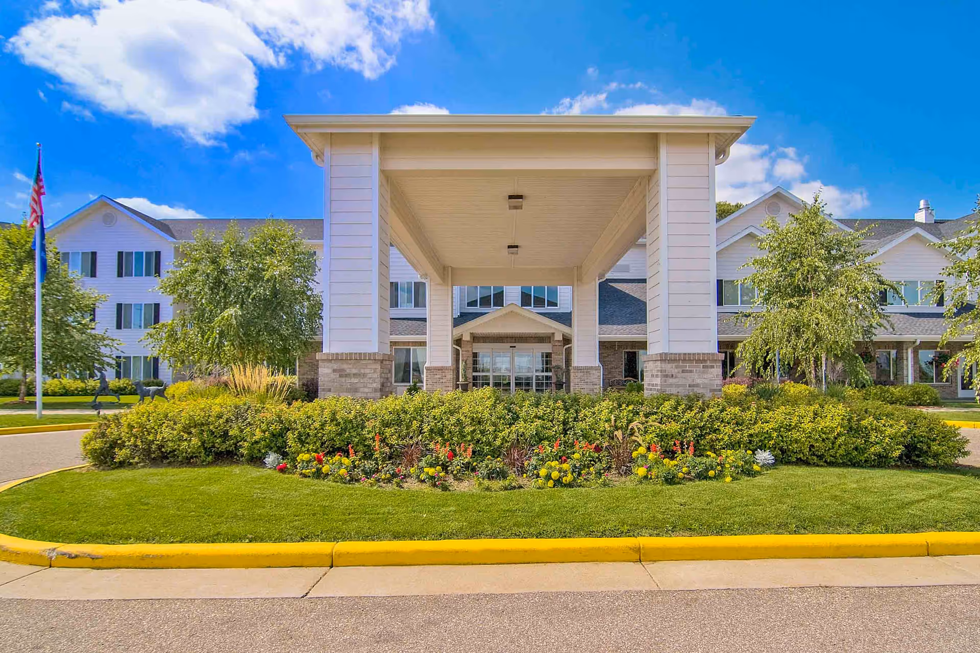 Front entrance of Oakwood Hills - Sky Active Living facility with a covered drop-off area, well-maintained landscaping including green bushes and colorful flowers, and a clear blue sky with some clouds.