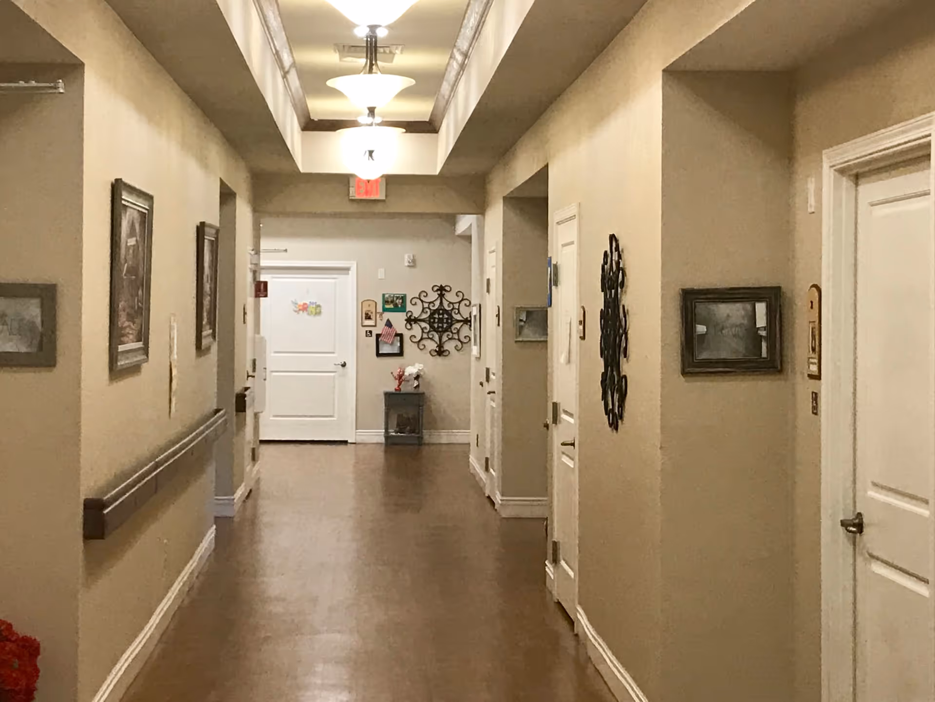 A well-lit hallway in a senior living facility with beige walls, wooden flooring, and white doors on both sides. The hallway is decorated with framed pictures and wall art, and there is a small table with flowers and decorations at the end near an exit door with an illuminated exit sign above it.